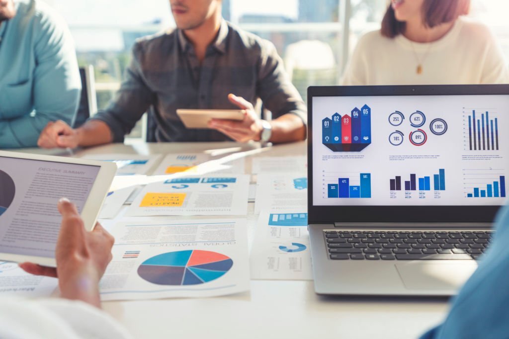 Group of people meeting with technology and paperwork. Close-up of digital tablet and laptop computer with market analysis data on the screen. The documents have financial or marketing figures, graphs, and charts on them. Multi-ethnic groups including Caucasian, Latin, and African American